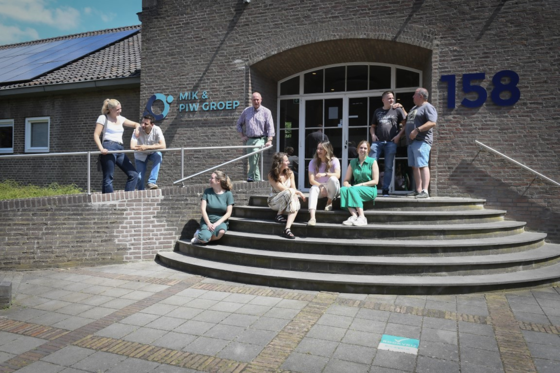 Negen medewerkers zijn met elkaar in gesprek op de trappen van ons kantoor in Beek.. Ze werken samen in de ondersteundende diensten van MIK & PIW Groep en ondersteunen sociaal werk en kinderopvangnvan MIK & PIW Groep in Beek. Ze zijn enthousiast met elkaar in gesprek. ij Knooppunt Informele Zorg.